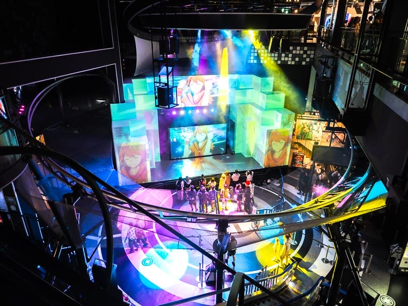 Looking down on an indoor concert, with a small crowd of people standing and watching some anime characters on big screens, with multicolored lights shining around