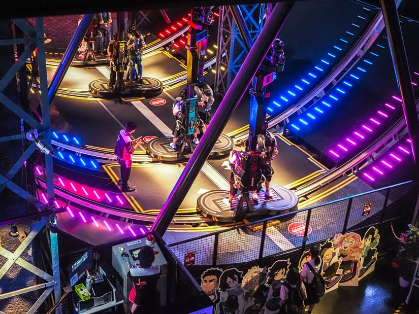Looking down on an amusement park ride inside a VR center, with some people getting strapped in to the ride and some purple and blue lights going up the circular track on either side of them