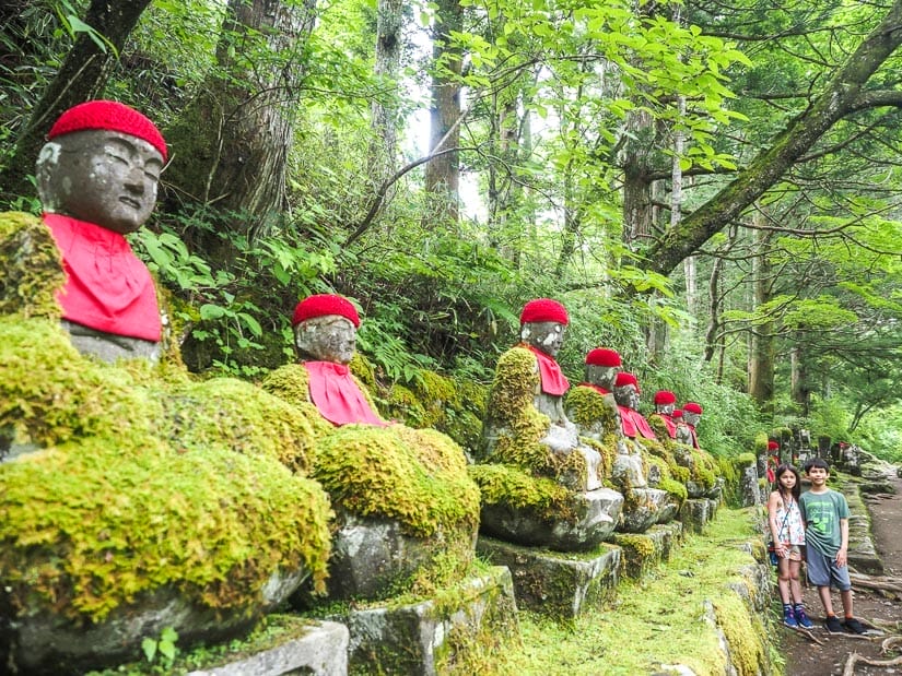 Two kids standing at the end of a low row of seated Buddha statues in the forest