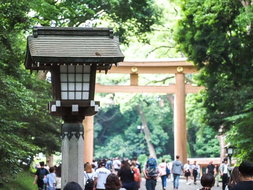 A stream of people walking below a tall wooden torii in a park and a traditional lamp sticking up in the foreground