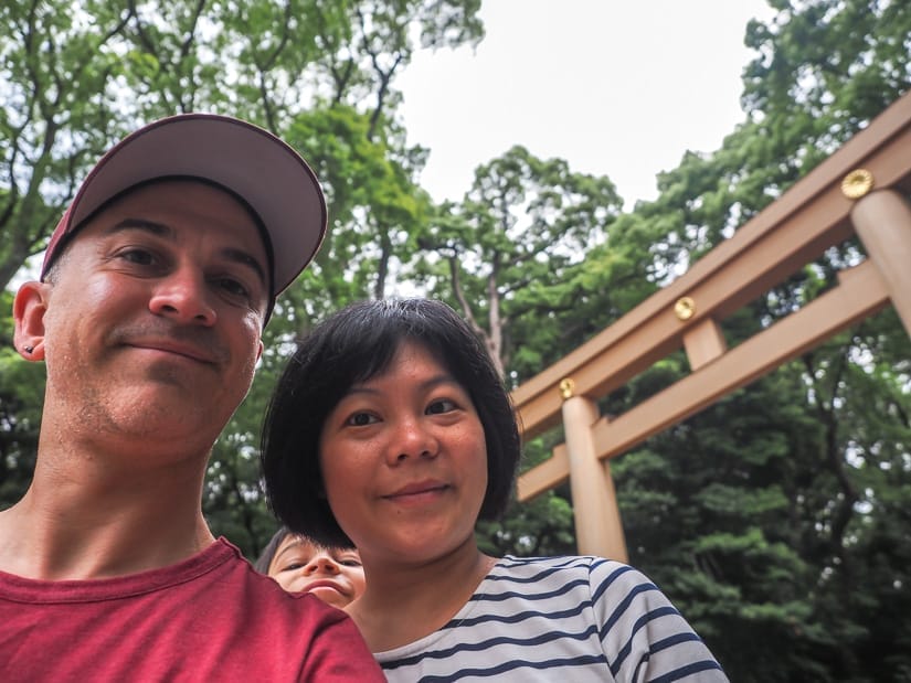 Nick Kembel and his wife Emily posing below a tall torii gate in Yoyogi Park in Tokyo, with their son photobombing between them