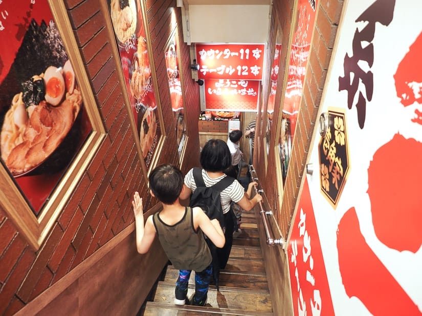 View looking down a stairwell, with a boy and woman descending the stairs, and Japanese signs and pictures of ramen on the walls on either side