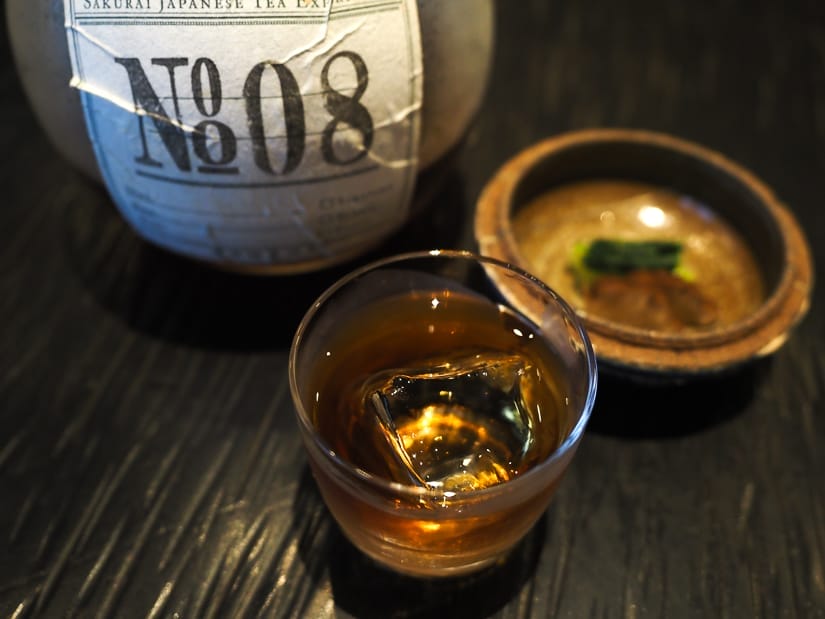 A small cocktail glass with dark brown liquid and ice cube in it, a small Japanese snack behind it, and a jar of liquor with label that says N808 behind it