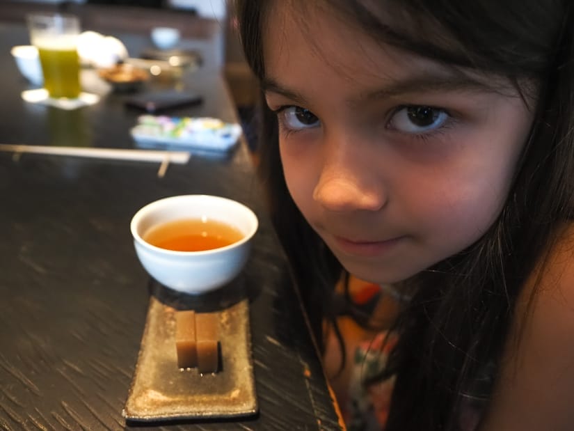 A young girl's face looking at the camera with a small tray with cup of tea and little Japanese dessert on the table in front of her