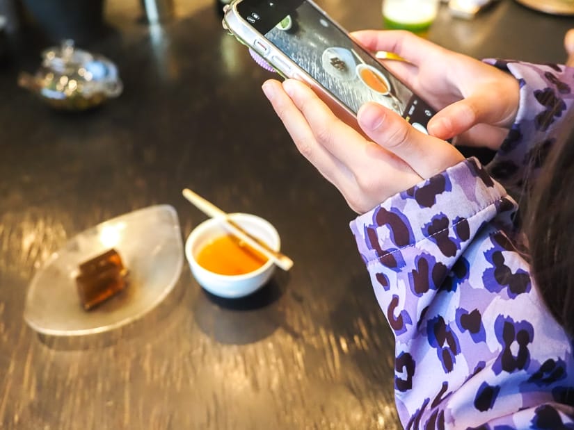 Close up of a girl's arms and hands as she holds a cell phone to take a photo of the tea cup and Japanese dessert in front of her