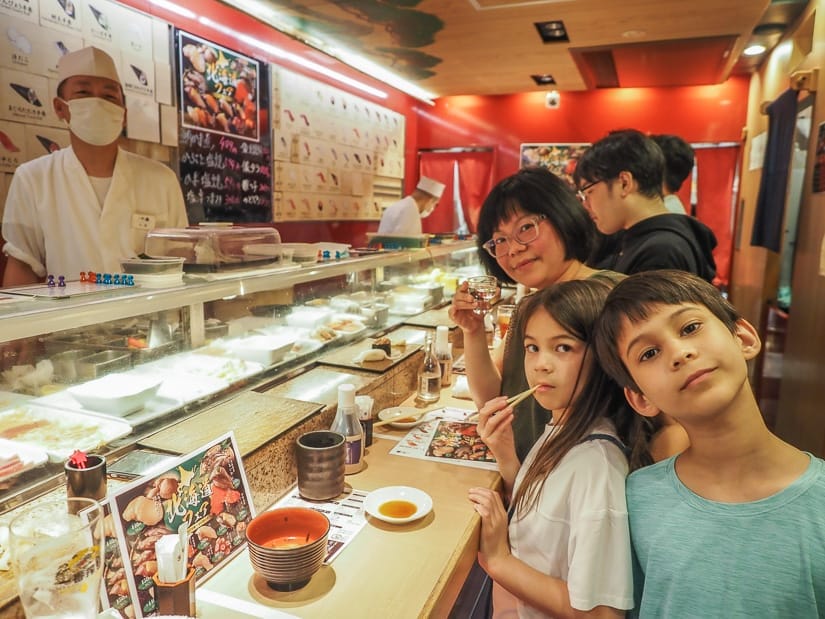 Two kids and a mom standing at a sushi bar, and the chef behind the bar is also looking at the camera
