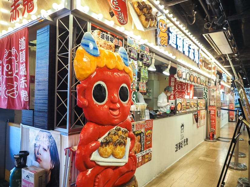 A takoyaki booth in a food court with weird red character eating takoyaki on the side