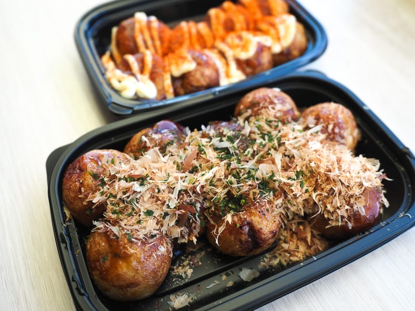 Two black trays of takoyaki, the one in the front with fish flakes and terriyaki sauce, the one in the back with creamy sauce and spicy powder