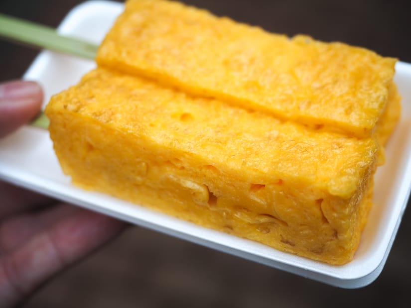 Close up of two rectangular pieces of Japanese tamogo (egg) on skewers, on a white take-away plate, held by a hand