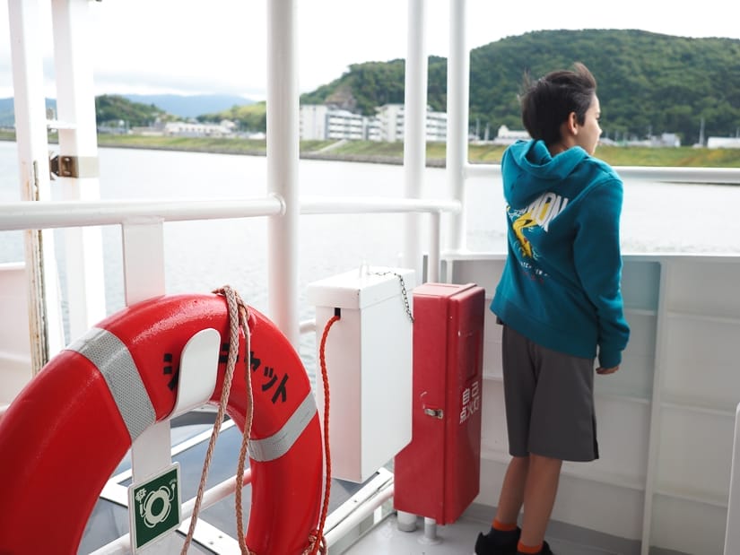 A young boy with Pokemon hoodie looking out over the side of a ferry, shot from behind, with an orange floating ring hanging on the railing to his left