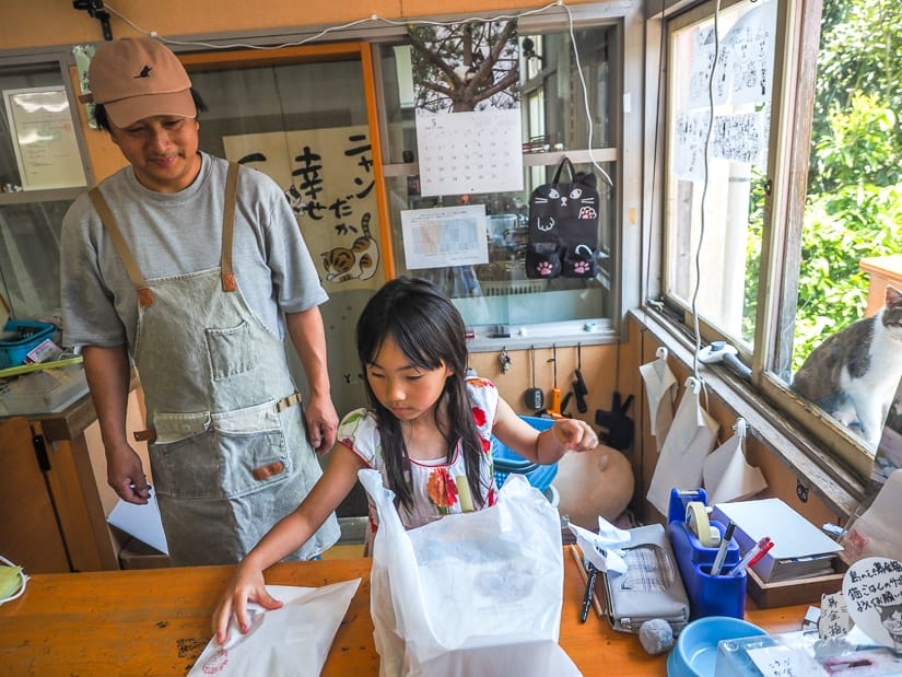 A young Japanese girl putting some purchases in a bag while her father watched from the left side, and a cat is visible outside the window on the right side.