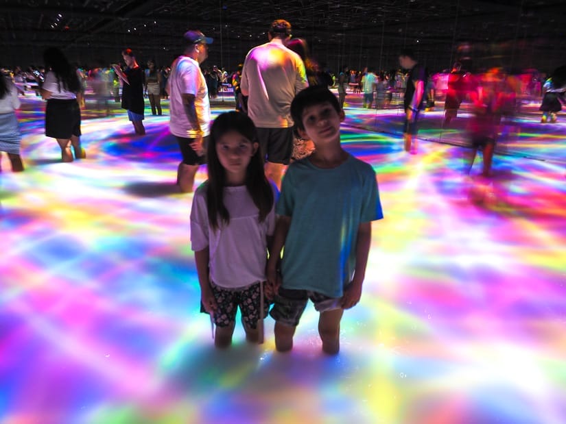 Two kids standing in a dark room filled to their knees with water and multicolored lights shining on the surface of the water