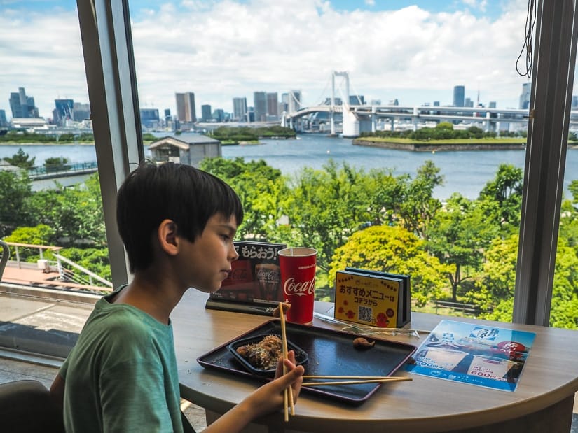 A boy eating takoyaki at a table next to a window with view of Tokyo harbor and bridge
