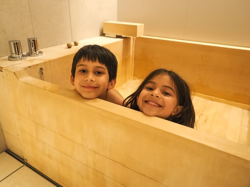 Two kids' heads poking up from a wooden, square shaped Japanese bath tub