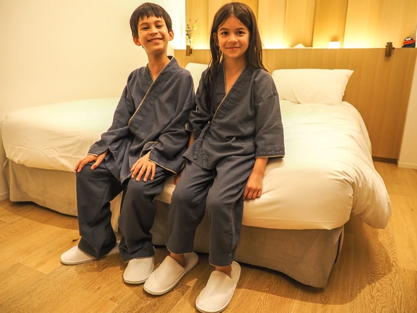A young boy and girl sitting on a bed in a hotel room in Tokyo, wearing matching blue traditional Japanese bath robes and white slippers