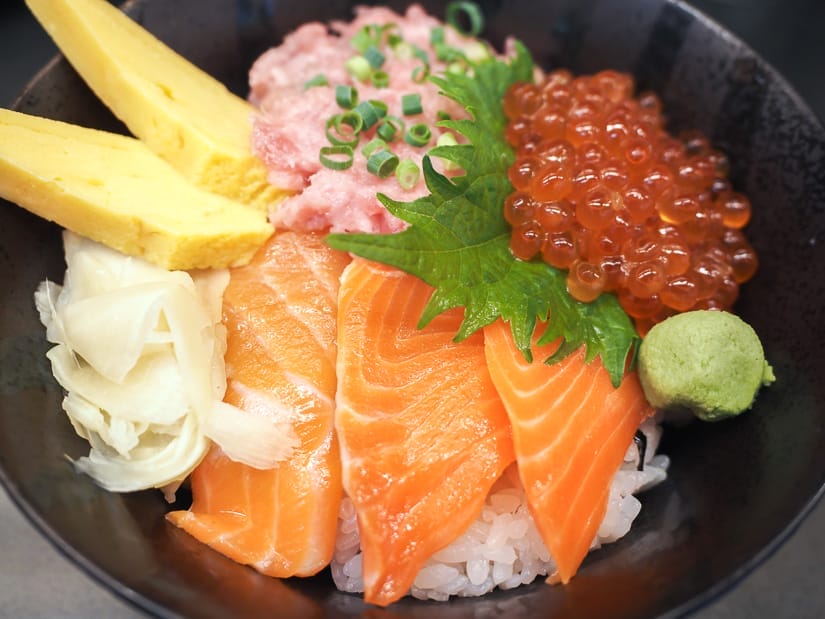 Close up of a bowl with raw salmon slices, salmon roe, mashed tuna, and tamago on rice