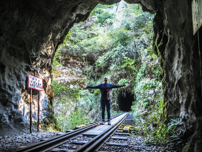 Nick Kembel standing in the entrance of a train tunnel with arms outstretched and train bridge leading to another tunnel behind him