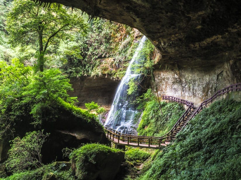 A wooden boardwalk trail passes below a waterfall pouring down from the top of the cave