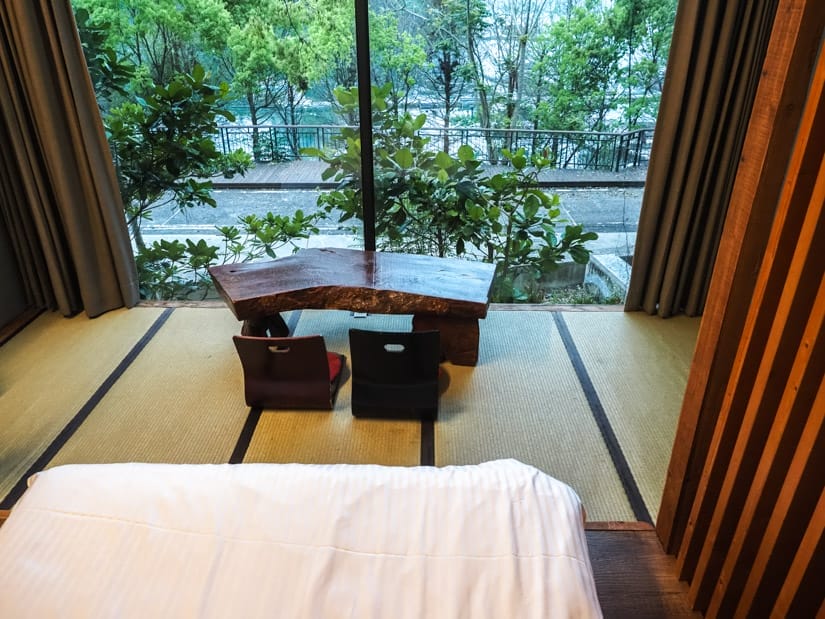 A photo shot from the bed of a hotel room, with the bottom of the bed in view, a low table with two stools on a Japanese-style tatami floor, and large windows facing a lakeside boardwalk with railing and trees