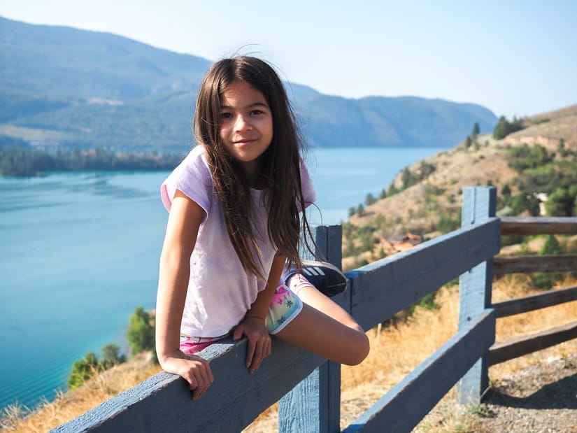 A young girl climbing over a wooden fence with an expansive view of Kalamalka Lake behind her