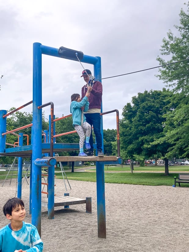 A father helping his daughter get onto a zip line in a playground