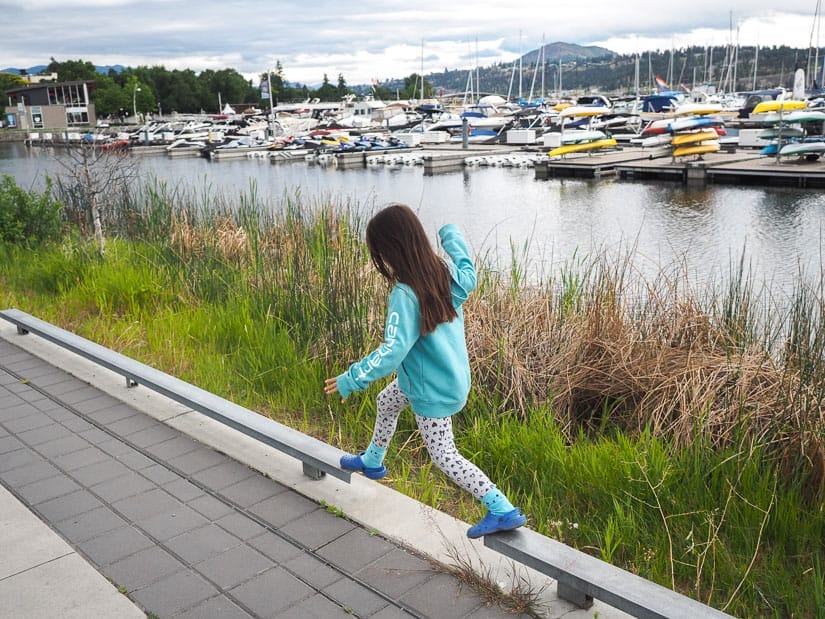 A young girl balancing her legs between two railings of a path beside a lake with many small docked boats