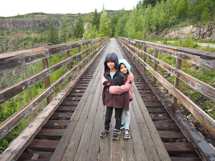 Two kids standing on a wooden trestle bridge, with the girl embracing the boy as they both face the camera