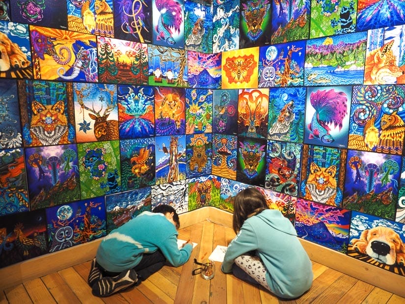 Two kids sitting on the floor coloring in front of a wall of colorful artworks
