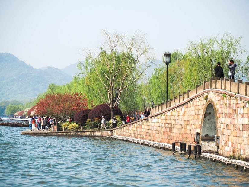 A small stone arched bridge on the side of a lake to a causeway with cherry blossoms
