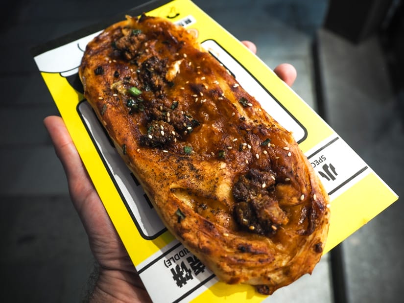 A hand holding up a flaky pastry with beef chunks and sauce on top, on top of a white and yellow wrapper