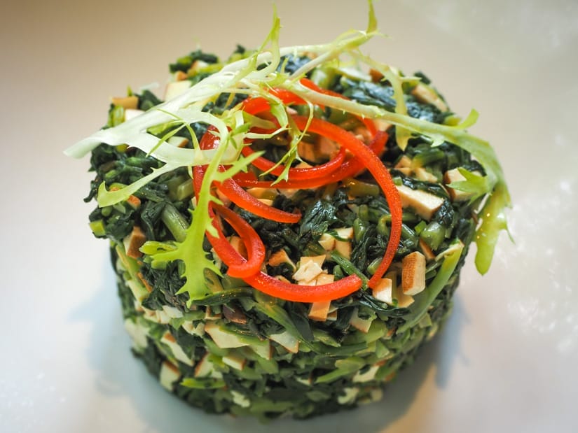 A muffin shaped mound of fried greens and tofu with red pepper swirls for garnish on a white plate
