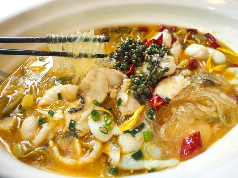 A large white plate with steamed fish in yellow soup and chopsticks holding up some noodles from it
