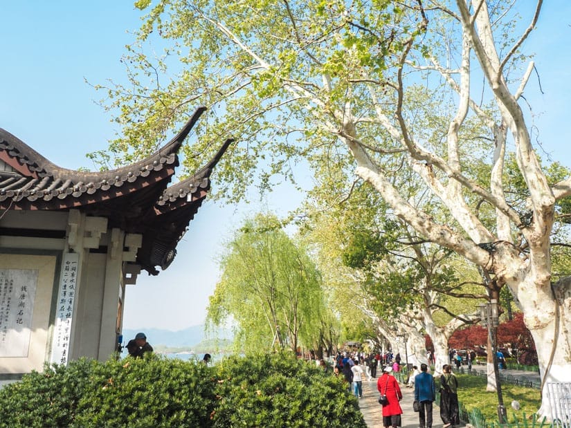 Hangzhou's West Lake, just barely visible between a pagoda-like structure on the left and some people walking on a lakeside path and some tall trees on the right