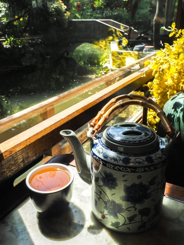 A pot of tea and filled tea cup on a table with yellow flowers in front of a window with view of pond and small bridge over it outside
