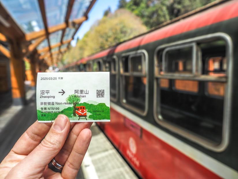 A hand holding up a ticket for the Zhaoping Line of Alishan Forest Railway with one of the red train cars parked at the train station platform behind