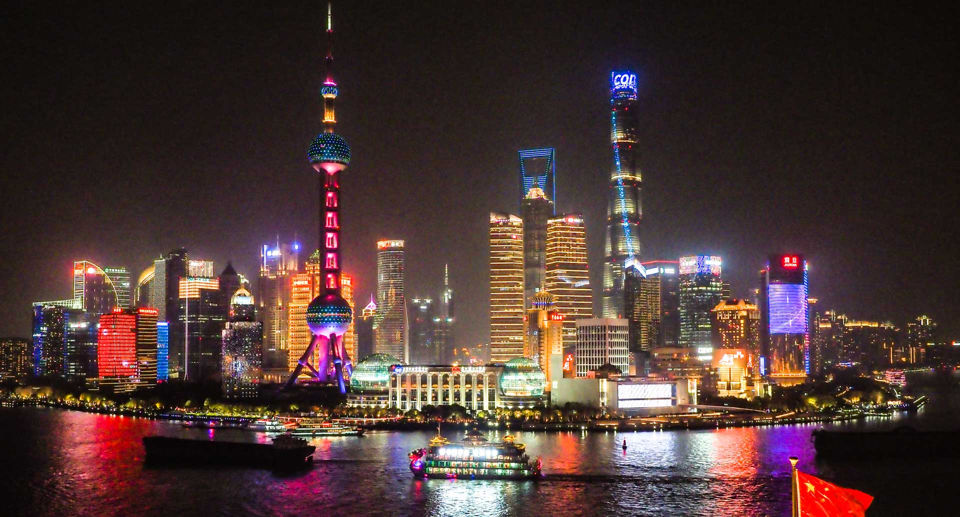 Pudong skyscrapers shot at night from across the river, with a flag of China flying at the bottom right in the foreground