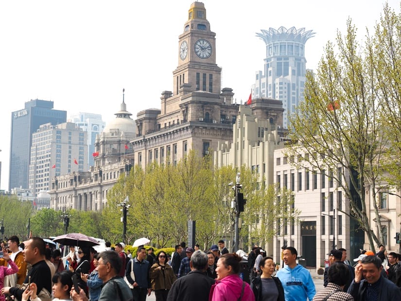 A crowd of people at the bottom with trees and a row of historic buildings along the Bund behind them
