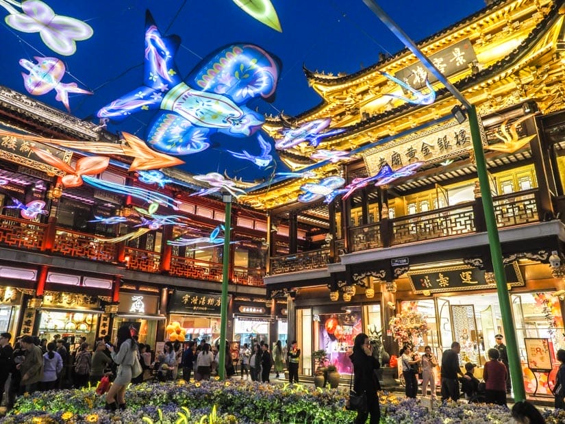 A night scene with a garden, traditional Chinese buildings around it, and some lit up kites forming a canopy above
