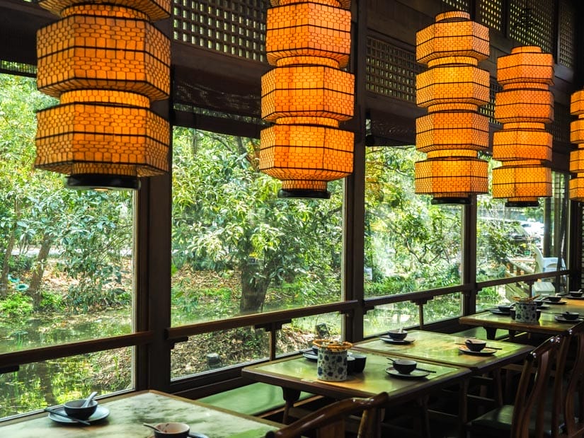 A restaurant dining room with tall lanterns above and window facing pond and foliage