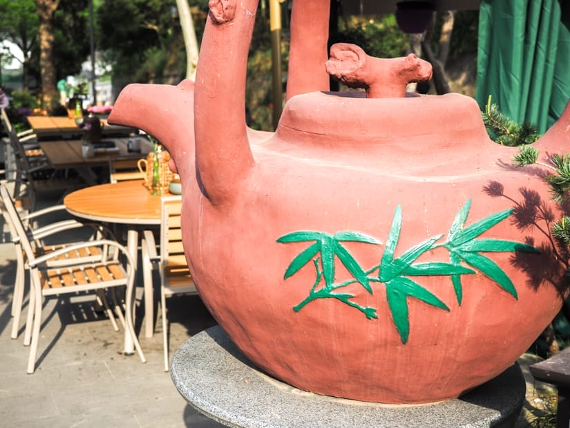 An oversized ceramic teapot with bamboo designs on it and teashop tables behind