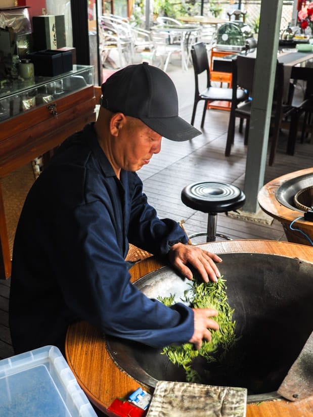 A Chinese man using his hands to move some Longjing tealeaves around in a large roasting pan