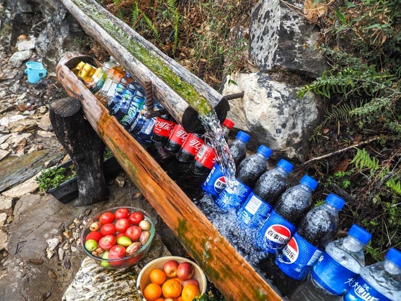 A stream of water running down a half bamboo tube onto some bottles of Coke and Pepsi, and some fruits for sale, and the side of a hiking trail