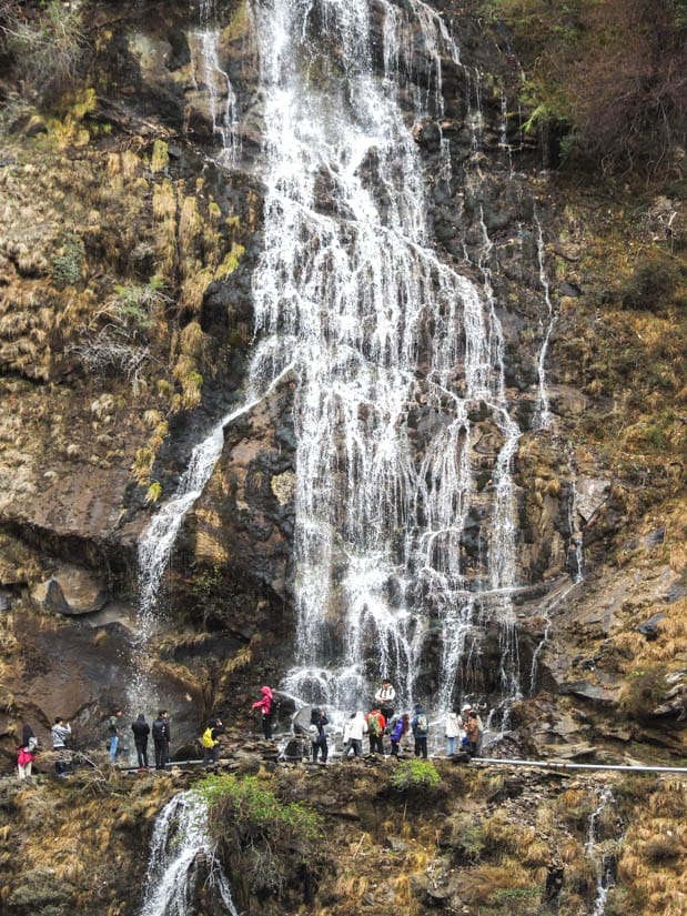 Zoomed in shot of a waterfall spilling down a cliff with some hikers on a trail in front of it