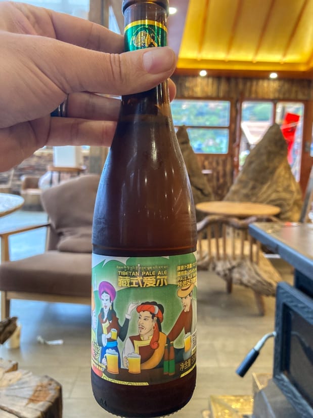 A hand holding up a bottle of Tibetan Pale Ale next to a wood burning stove inside a cafe