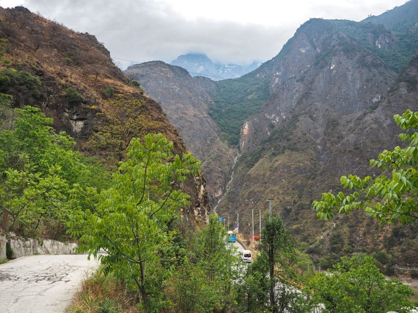 Looking down a small paved road at a highway with buses going by and scenery of Tiger Leaping Gorge beyond