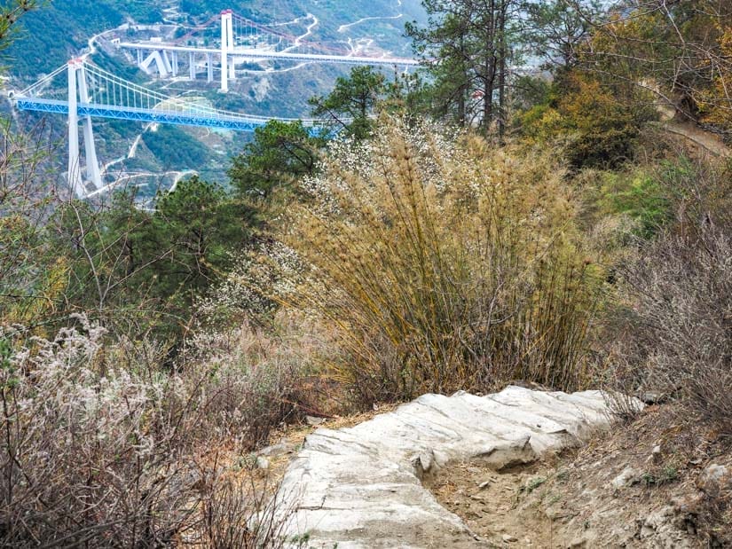 Looking down a narrow hiking path, with a blue bridge behind and far below in the gorge