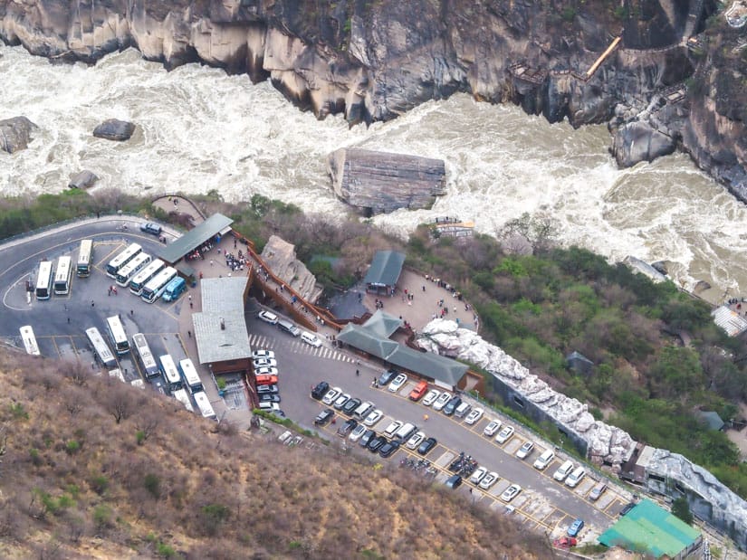 Looking down on a parking lot filled with cars and tour buses next to the river in Tiger Leaping Gorge, with huge stone in the middle of the river